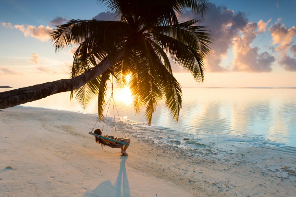 Person sitting on a palm tree swing on a Maldives beach at sunset with calm ocean reflections