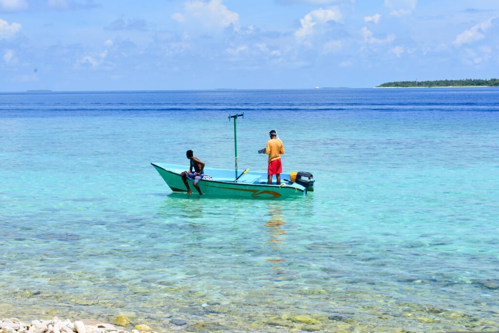 Two Maldivian fishermen on a traditional green fishing boat in crystal clear turquoise water near a local island.